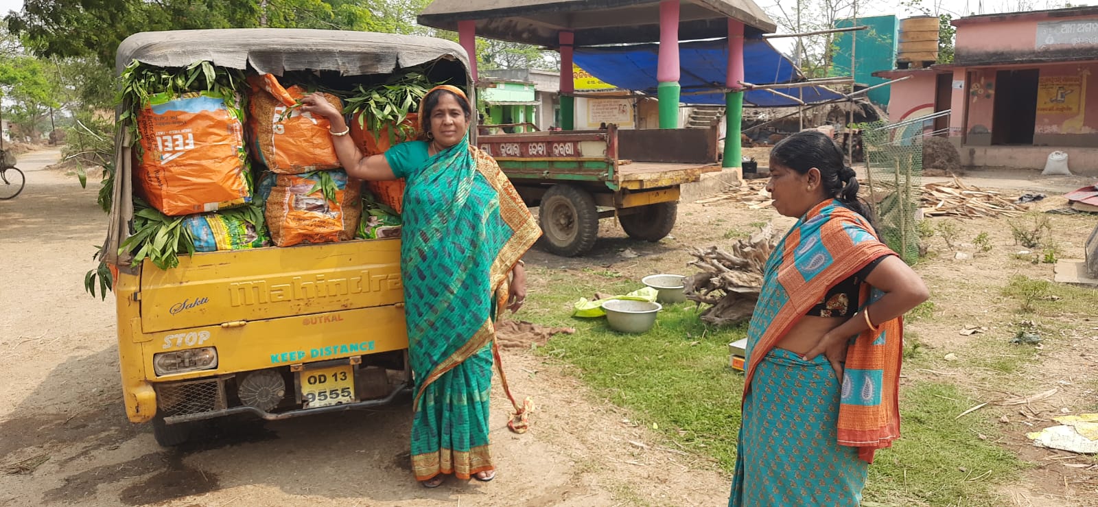 Indian woman in sari stands next to a truck filled with produce while another Indian woman looks on
